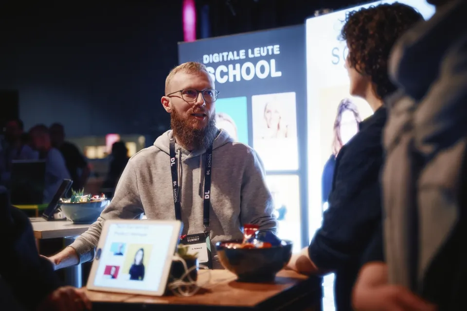 DL-School-Stand 2025: Berater mit DL-School-Lanyard im Gespräch, Tablet zeigt „Tech-Karriere als Product Manager“, Banner „Digitale Leute School“